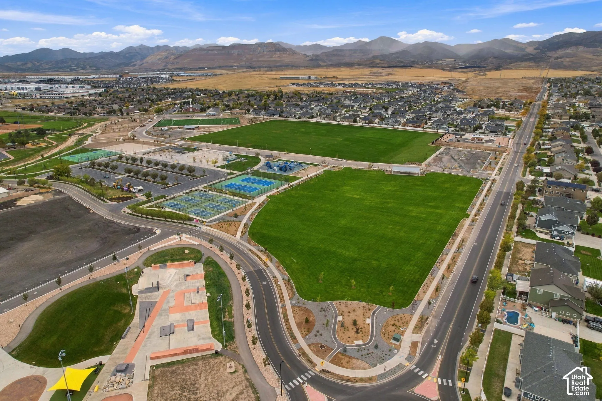 Aerial perspective of the neighborhood featuring expansive green space, multiple tennis courts, a paved skate park, and a mountain backdrop