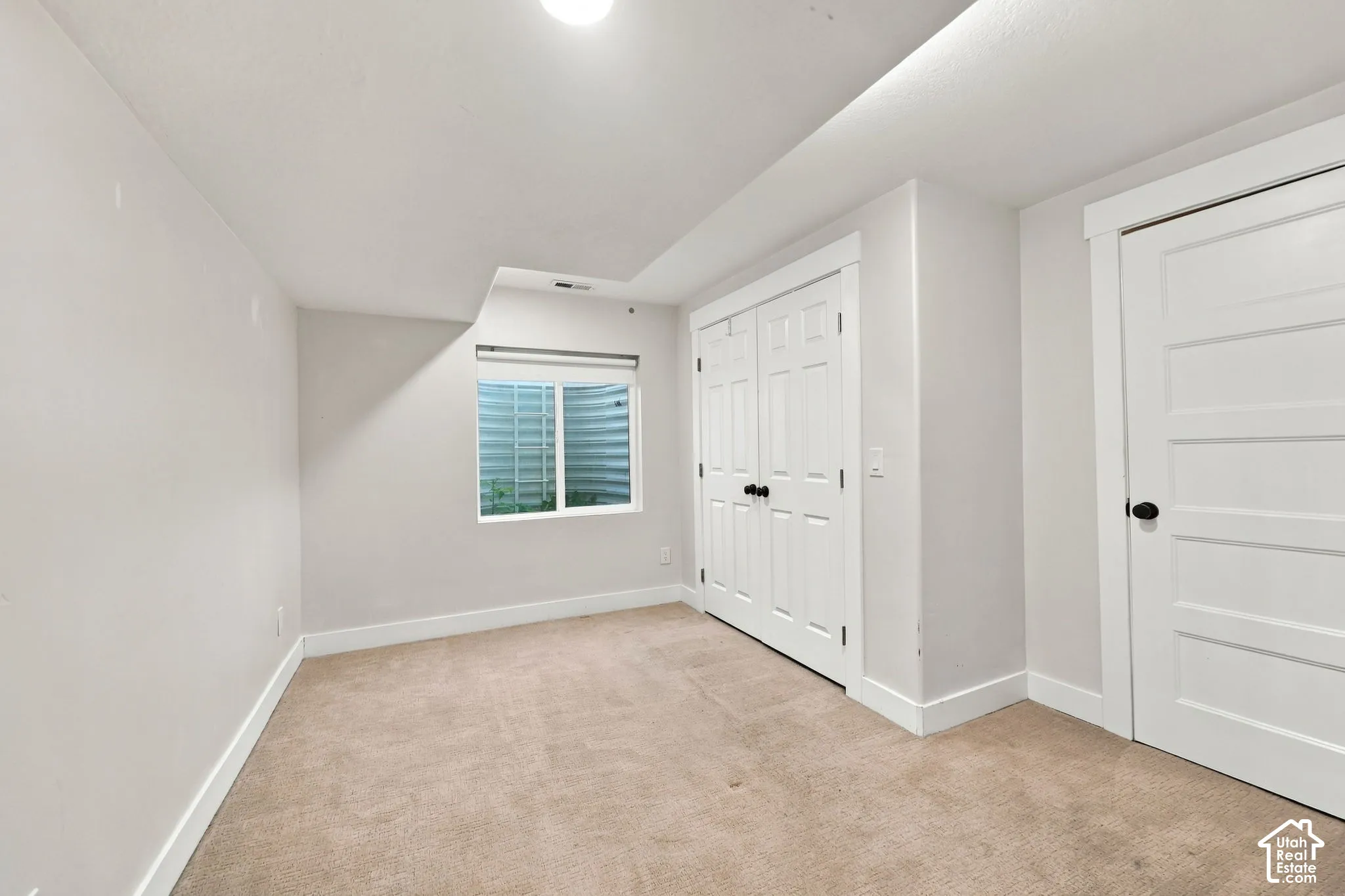 Neutral-toned interior space featuring carpeted flooring, a recessed window with a window well, double-panel closet doors, and a single-panel door with matte black hardware