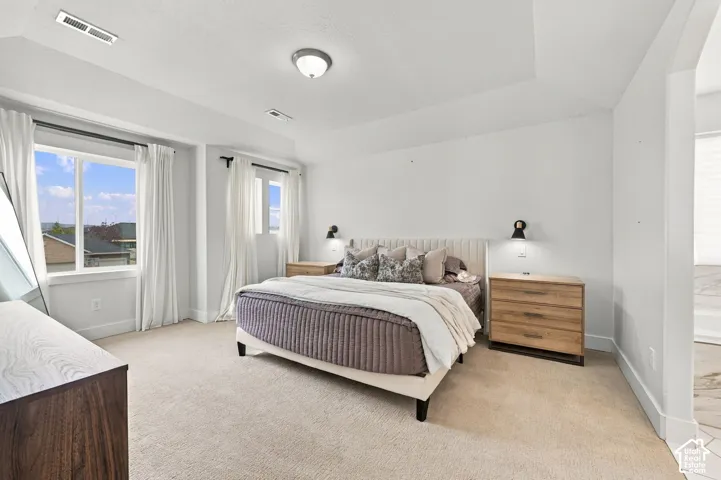 Tray ceiling with central flush-mount lighting, wall-mounted side sconces, multiple windows with neutral-toned trim, and wall-to-wall carpeting