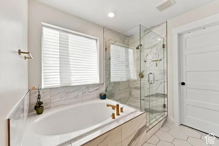 Oval soaking tub with brass deck-mounted faucet, walk-in shower with frameless glass enclosure, stone-finish tile surround, and hexagonal floor tiling