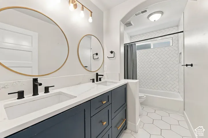 Dual-sink vanity with matte black fixtures, white stone-finish countertops, herringbone-patterned wall tile, and hexagonal floor tile