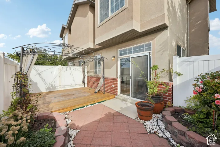 Exterior space featuring a wood deck, red-toned paved patio, brick accents, and a white privacy fence with built-in stone garden borders