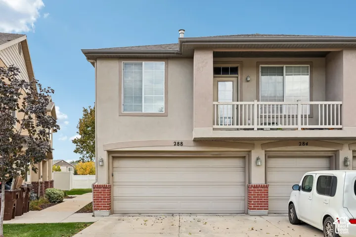 Stucco exterior facade featuring a covered upper-level balcony with railing, integrated two-car garage, and decorative brick accents at the base
