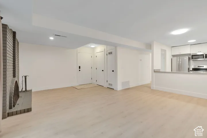 Open-concept interior featuring light wood-finish flooring, a full-height brick fireplace, recessed lighting, and a kitchen equipped with stainless steel appliances and white cabinetry