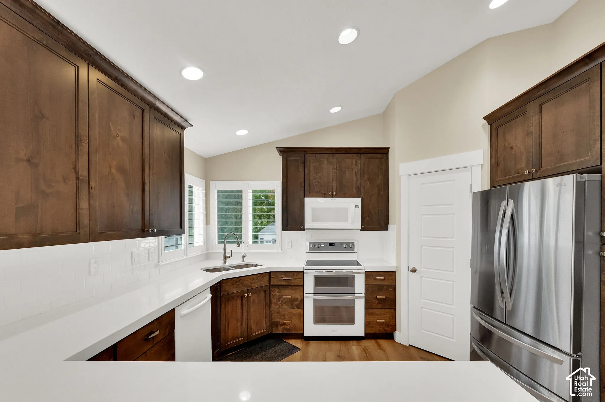 Kitchen featuring dark wood cabinetry, white countertops, white subway tile backsplash, stainless steel appliances, and recessed lighting under a vaulted ceiling