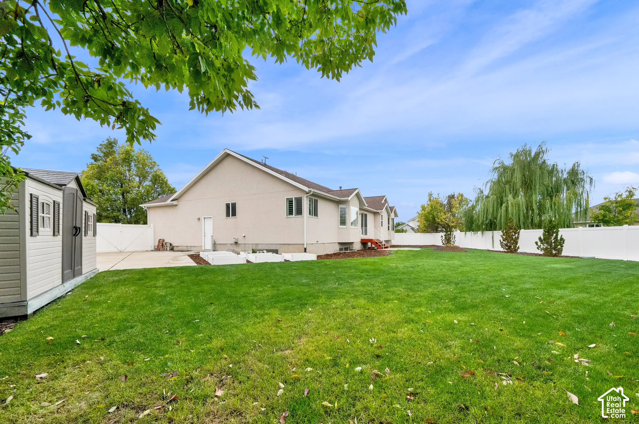 Rear exterior featuring a stucco facade, expansive lawn, white vinyl fencing, concrete patio, and a detached storage structure