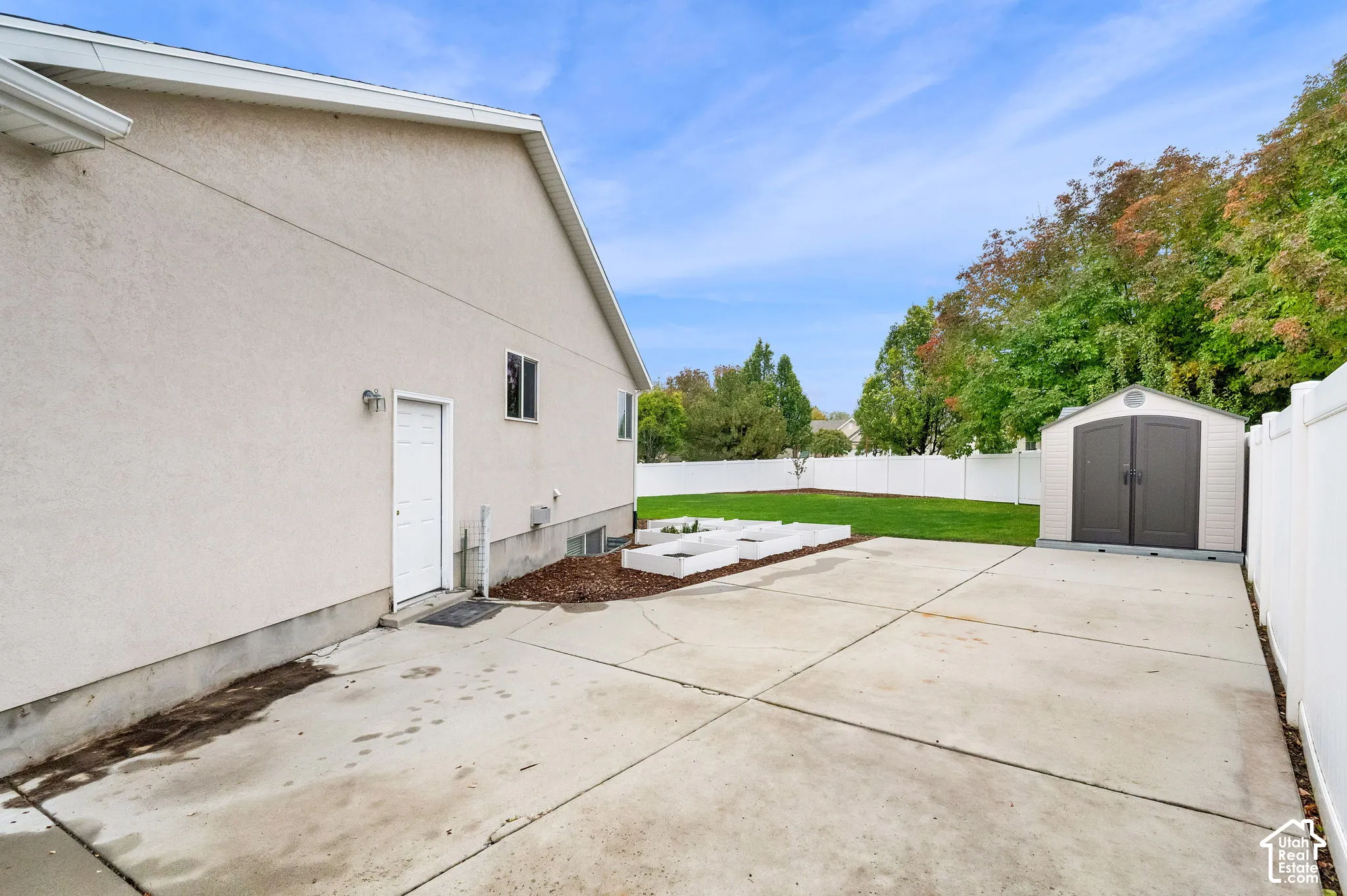 Backyard featuring a concrete parking pad, white vinyl perimeter fencing, raised garden beds, and a detached storage shed