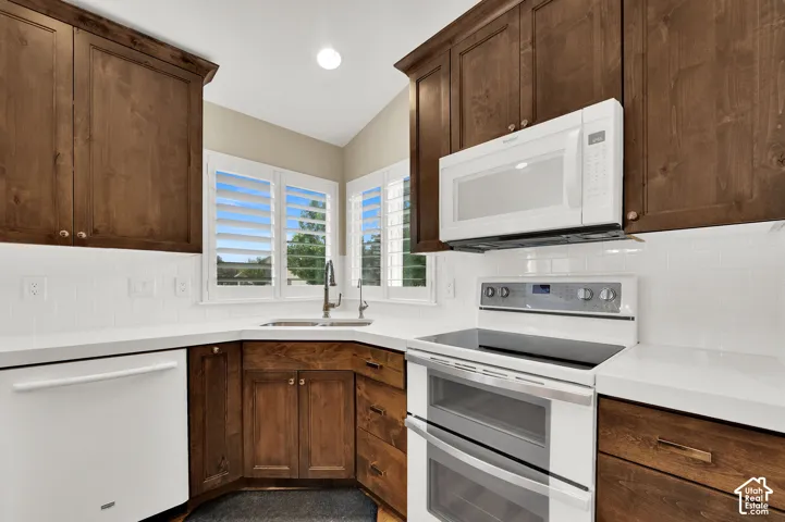 Stained wood cabinetry, white subway tile backsplash, solid-surface countertops, white appliances, and plantation shutters
