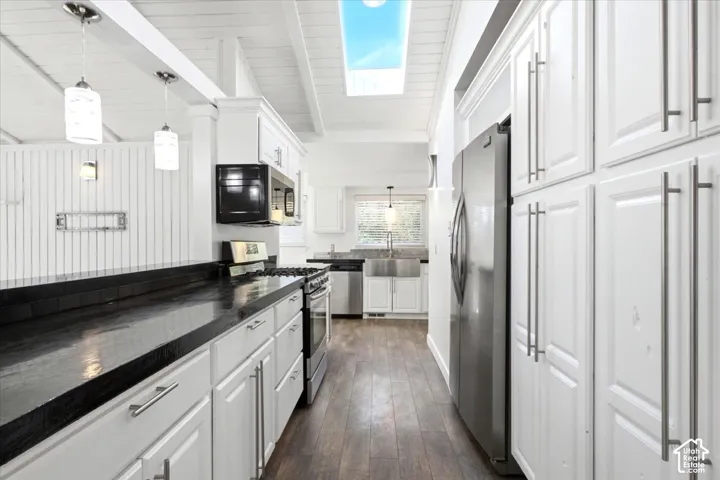 Kitchen featuring white shaker-style cabinetry, stainless steel appliances, dark countertops, wood-finish flooring, and a skylight