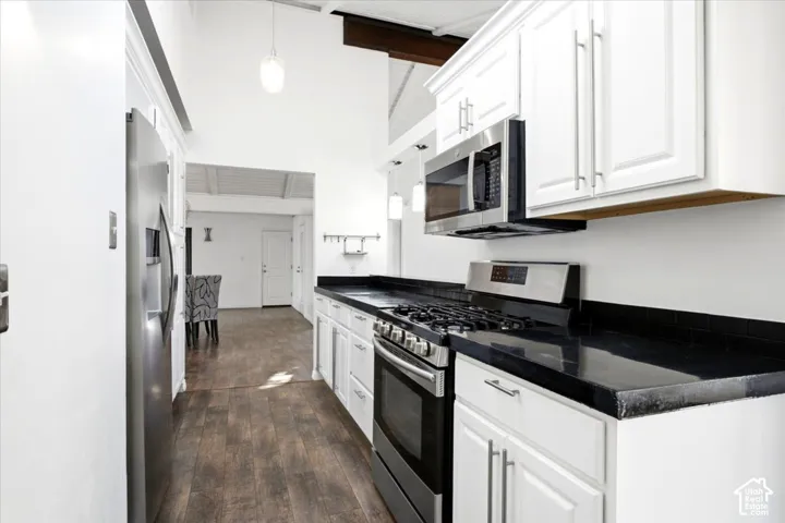 Kitchen featuring white Shaker-style cabinetry, dark stone-finish countertops, stainless steel appliances, wood-finish flooring, and vaulted ceilings with exposed structural beams