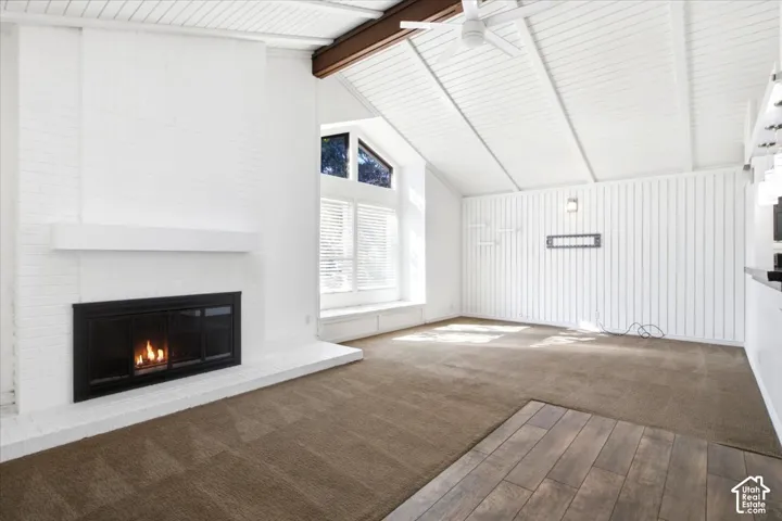 Vaulted ceiling with exposed wood beam, painted brick fireplace with mantel, vertical wood wall paneling, built-in window seat, and large geometric windows
