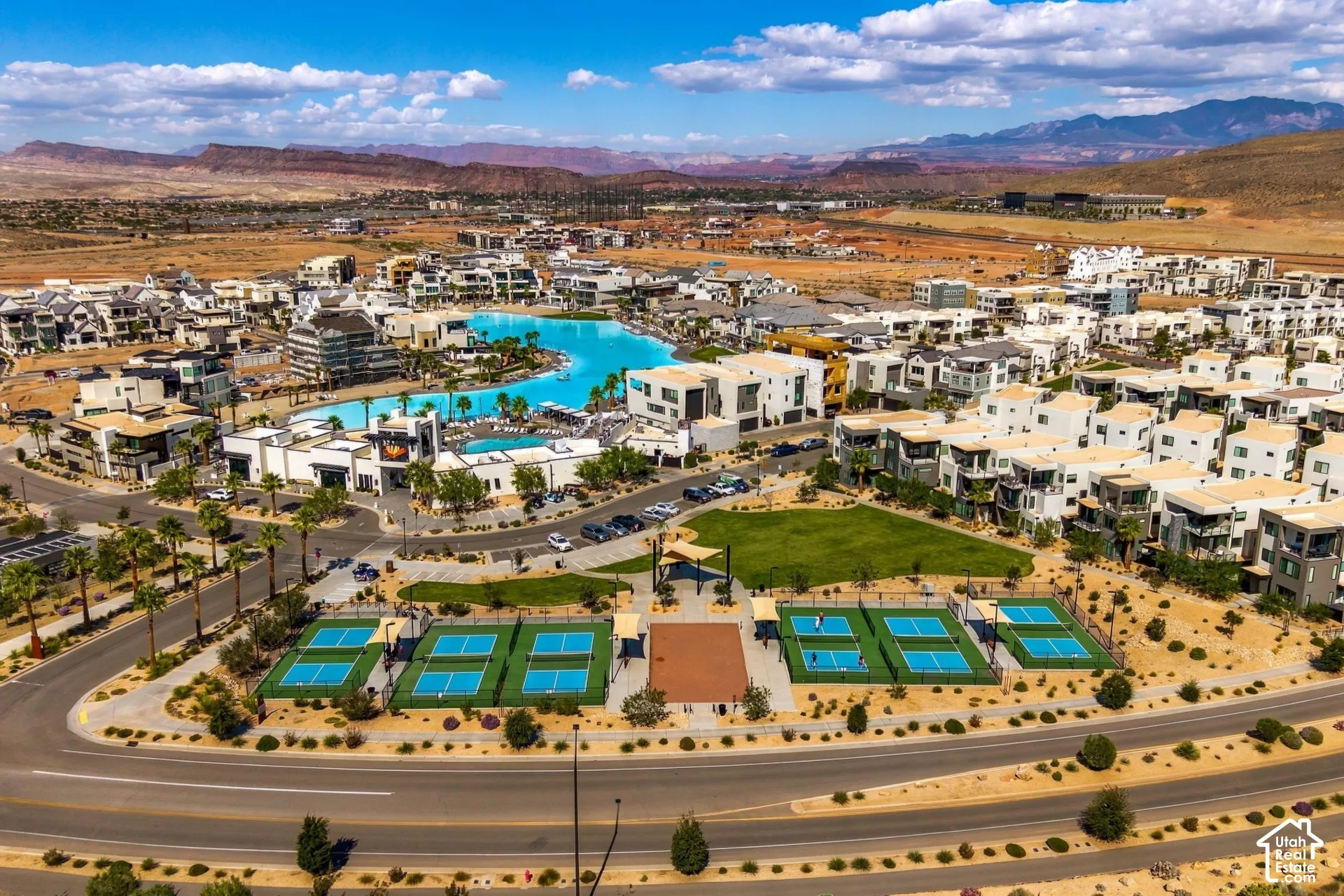 Aerial perspective of a master-planned community featuring a large turquoise lagoon, multiple pickleball courts, expansive green space, and a mountainous desert backdrop