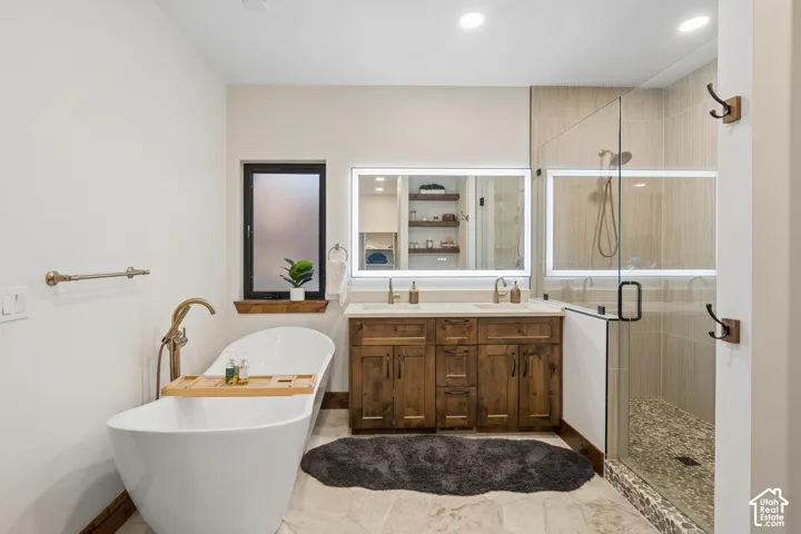 Freestanding soaking tub with a brass-finish floor-mounted faucet, dual vanity with shaker-style wood cabinetry and undermount sinks, illuminated mirrors, and a glass-enclosed shower with pebble-tile flooring