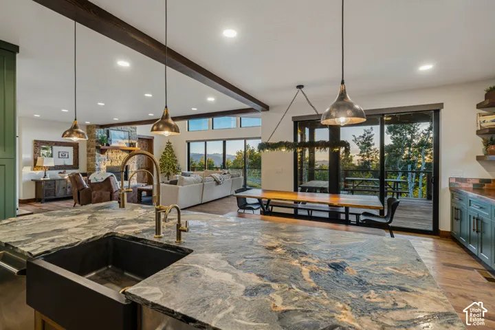Spacious open-concept living area featuring exposed ceiling beams, a built-in stone fireplace, large sliding glass doors, and a kitchen island with a farmhouse sink and brass-finish faucet
