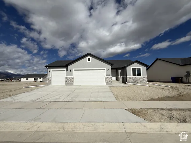 Single-level exterior featuring grey stucco siding, stone veneer accents, a shingled roof, a three-car garage, and a concrete driveway