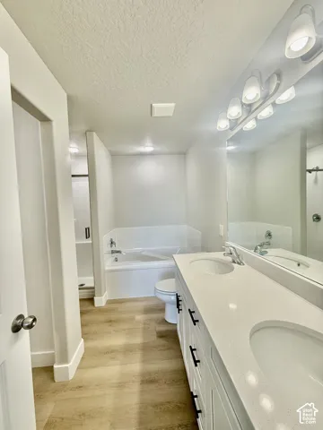 Dual-sink vanity with white cabinetry, light-toned stone-finish countertop, wood-finish flooring, drop-in bathtub with tiled surround, and separate glass-enclosed shower