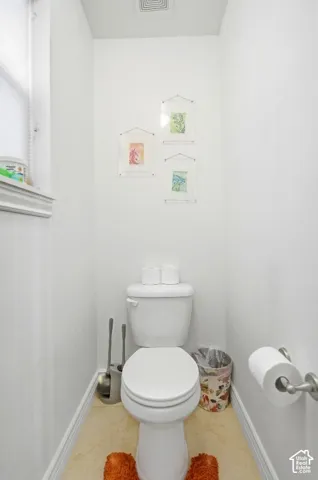 Restroom featuring a white porcelain commode, neutral-toned flooring, a window with horizontal blinds, and a wall-mounted toilet paper holder
