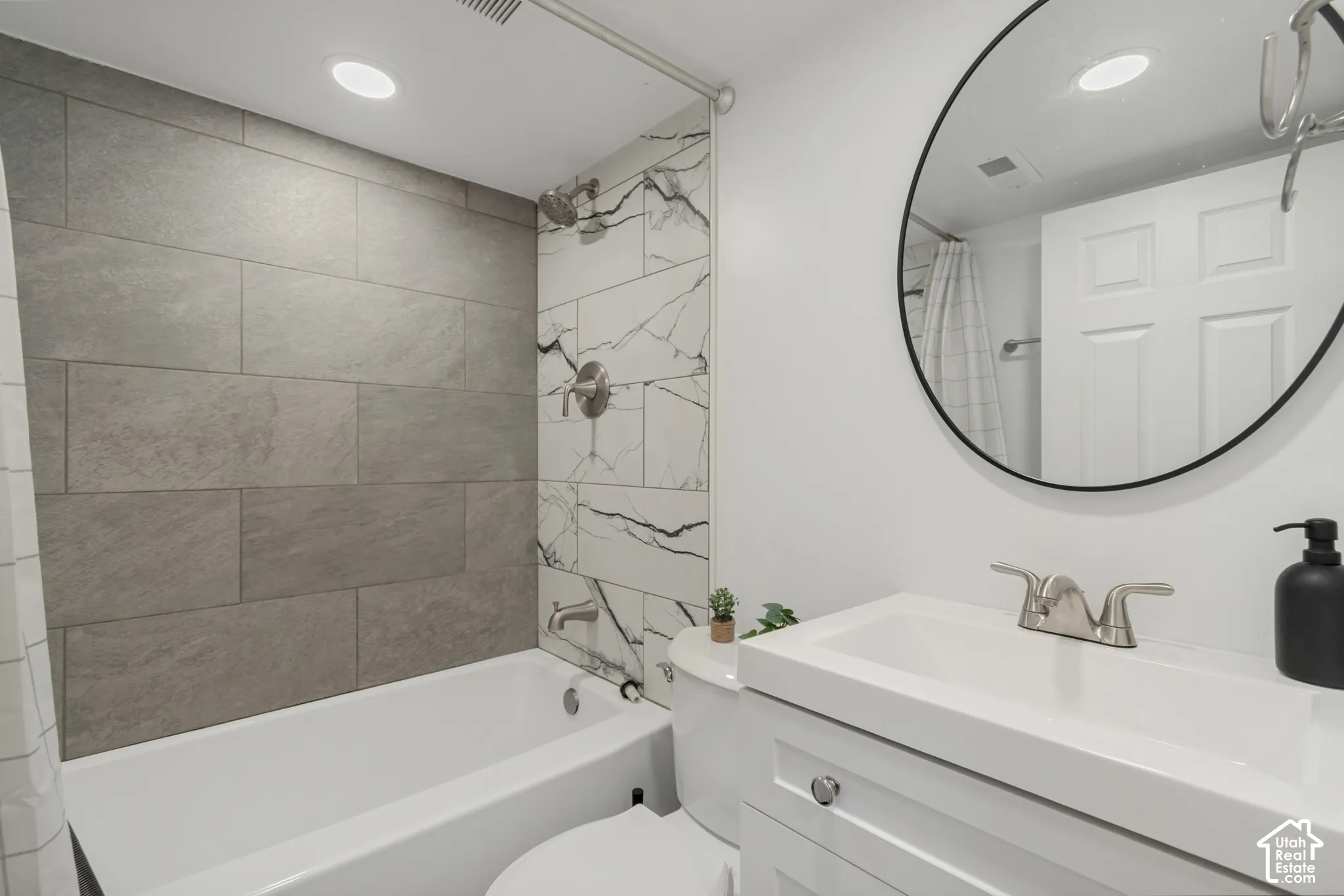 Bathroom featuring a vanity with an integrated rectangular sink, large-format grey tile tub surround, accent tile wall with marble-style veining, recessed lighting, and a circular wall-mounted mirror with a black frame