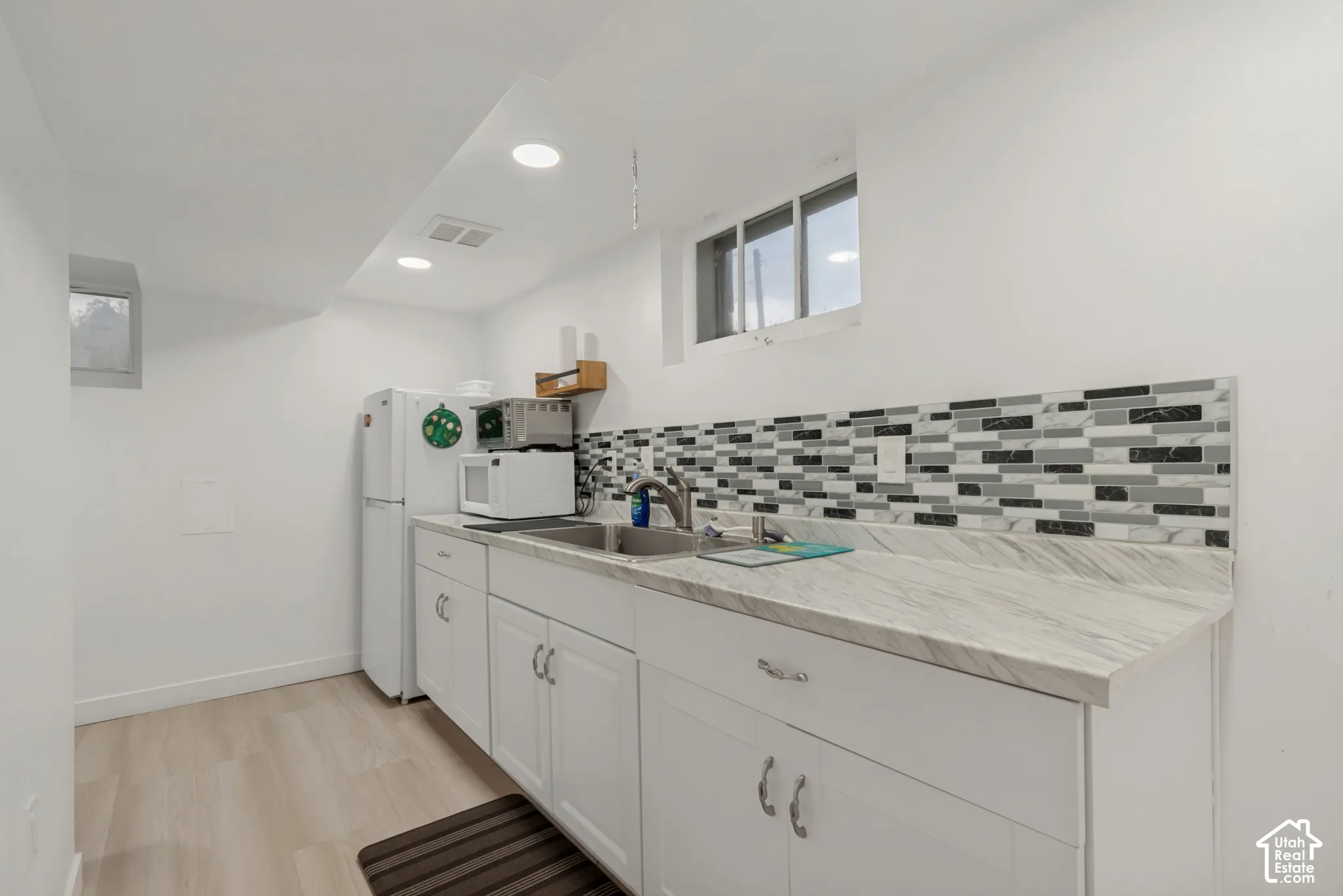 Kitchenette featuring white cabinetry, stone-patterned laminate countertops, multi-tonal linear tile backsplash, stainless steel dual-basin sink with arched faucet, and recessed lighting