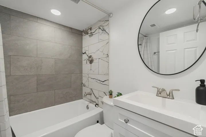 Bathroom featuring a vanity with an integrated rectangular sink, large-format grey tile tub surround, accent tile wall with marble-style veining, recessed lighting, and a circular wall-mounted mirror with a black frame
