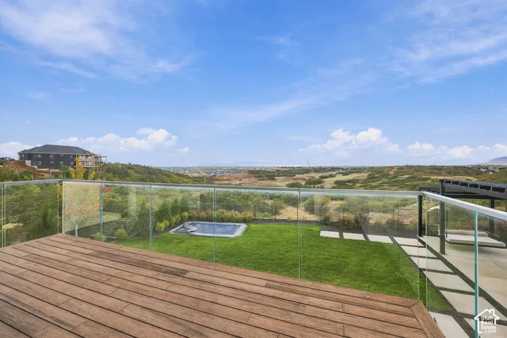 Elevated deck featuring wood-finish flooring and a frameless glass railing overlooking an expansive yard with a manicured lawn, an in-ground spa, and paved hardscaping