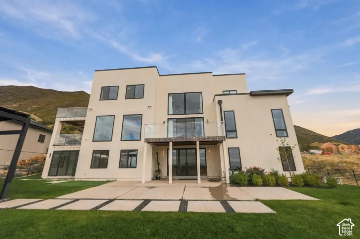 Modern multi-story exterior featuring smooth stucco siding, large fixed and casement windows with dark frames, elevated glass-railing balconies, and a paved concrete patio with stone-accented grass dividers