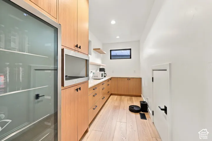 Custom wood-finish cabinetry with matte black hardware, integrated glass-front appliance, light-toned wood-finish flooring, floating shelves, and recessed ceiling lighting