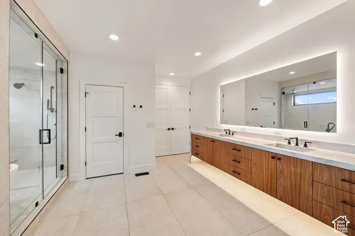 Floating wood-finish vanity with dual integrated sinks and stone-finish countertop, expansive wall-to-wall LED-backlit mirror, large walk-in glass-enclosed shower with tile surround, and matte black hardware fixtures