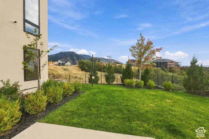 Landscaped backyard featuring a manicured lawn, dark gravel perimeter beds, ornamental shrubs, metal perimeter fencing, and a concrete patio
