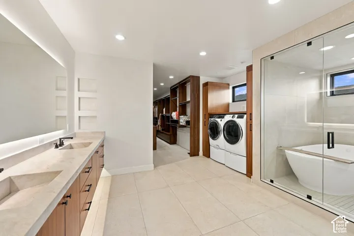 Double vanity with stone countertop and integrated sinks, glass-enclosed soaking tub, custom wood-finish cabinetry, recessed lighting, and built-in laundry appliances