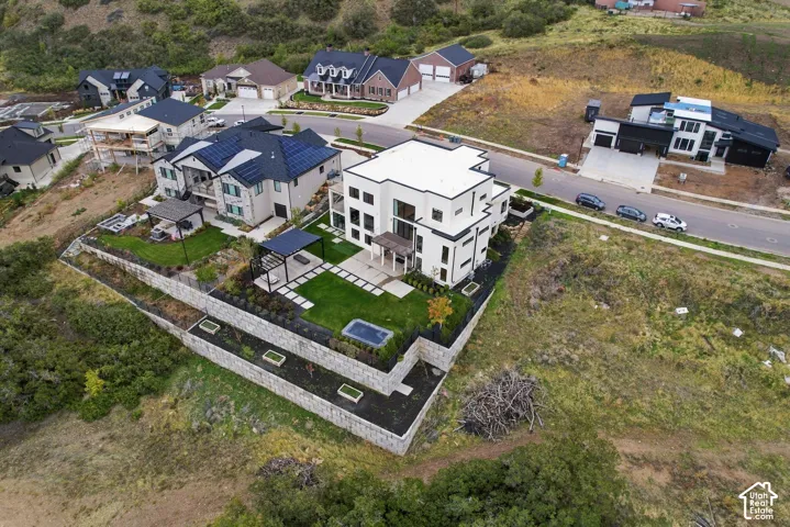 Aerial perspective of a multi-level residence featuring tiered stone retaining walls, a landscaped lawn, a concrete patio, and a covered pergola structure