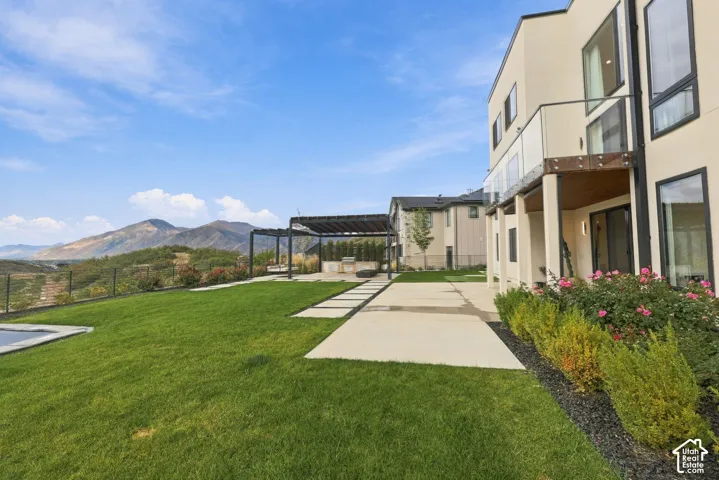 Rear exterior featuring a tiered concrete patio, pergola-covered outdoor kitchen, glass-railing balcony, and mountain scenery