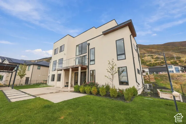 Contemporary stucco exterior featuring a tiered balcony with a glass railing, large rectangular windows with dark frames, a concrete patio, and landscaped ground cover