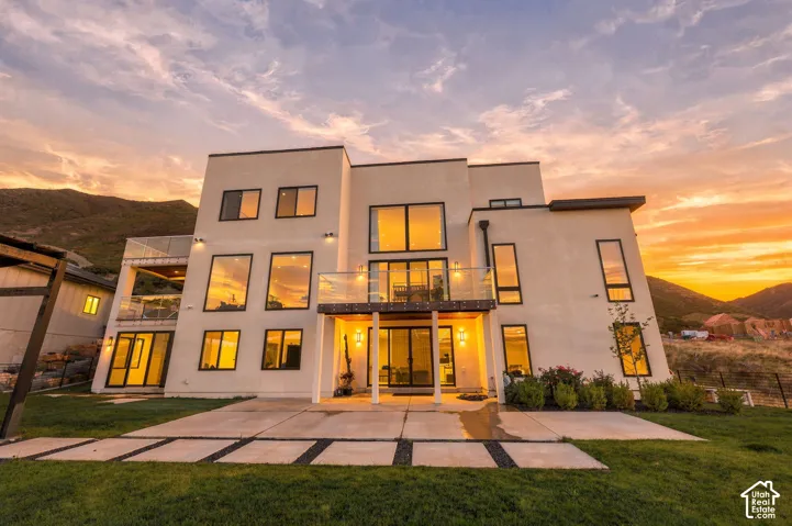 Modern stucco exterior featuring expansive glass windows, multi-level glass-railed balconies, and a concrete patio set against a mountain backdrop