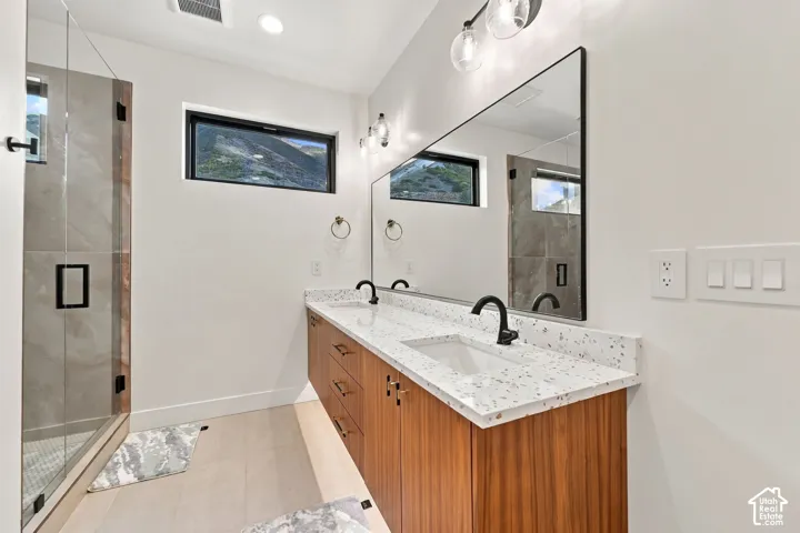 Modern dual-sink vanity featuring wood-finish cabinetry, speckled solid-surface countertops, matte black faucets, and a frameless glass-enclosed shower with a walk-in configuration
