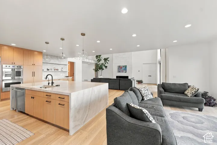 Kitchen featuring a waterfall-edge island, light wood-finish cabinetry, built-in stainless steel double ovens, recessed lighting, and wood-finish flooring