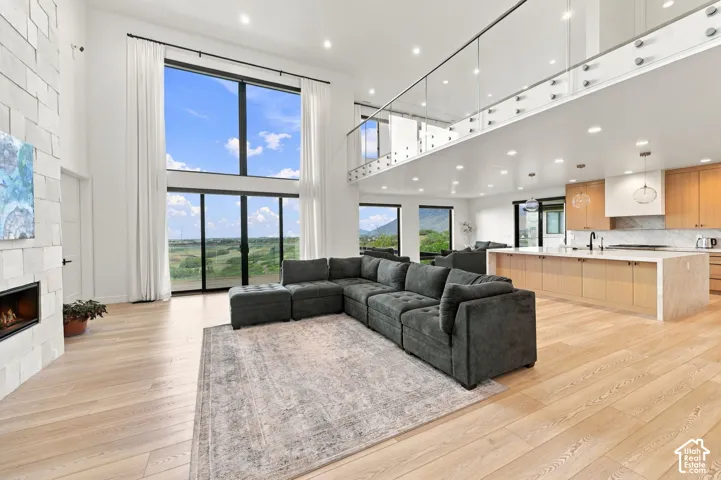 Double-height ceilings with floor-to-ceiling glass windows, stacked-stone fireplace, wood-finish flooring, glass-panel mezzanine railing, and expansive kitchen island with stone-finish waterfall edge