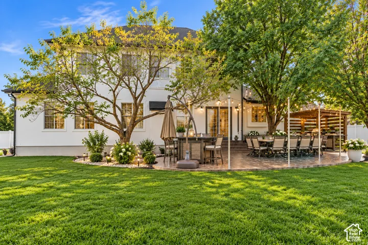 Rear exterior featuring a stucco facade, stone-paver patio, wood-slat pergola, and manicured landscaping with mature trees
