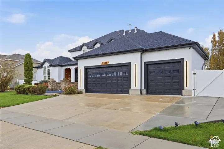 Exterior featuring a light-colored stucco facade, dark shingled roof, attached three-car garage with black paneled doors, vertical wall-mounted lighting, and concrete driveway