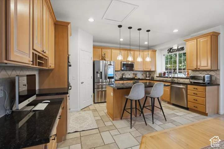 Kitchen featuring light wood-finish cabinetry, dark stone countertops, stainless steel appliances, central island with pendant lighting, and stone tile flooring