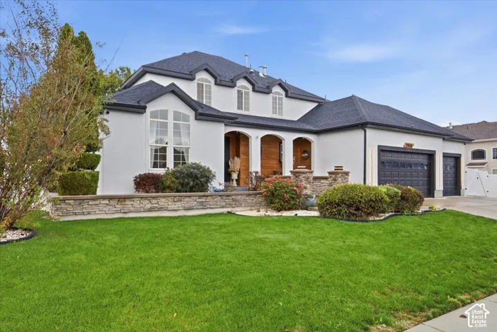 Two-story exterior featuring a white stucco facade, charcoal-toned shingled roof, stacked stone retaining wall, arched covered entryway with wood paneling, and a three-car garage