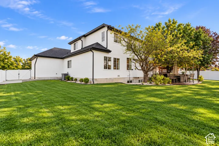 Two-story exterior featuring light-colored stucco siding, a dark shingle roof, a manicured lawn, perimeter fencing, and mature trees