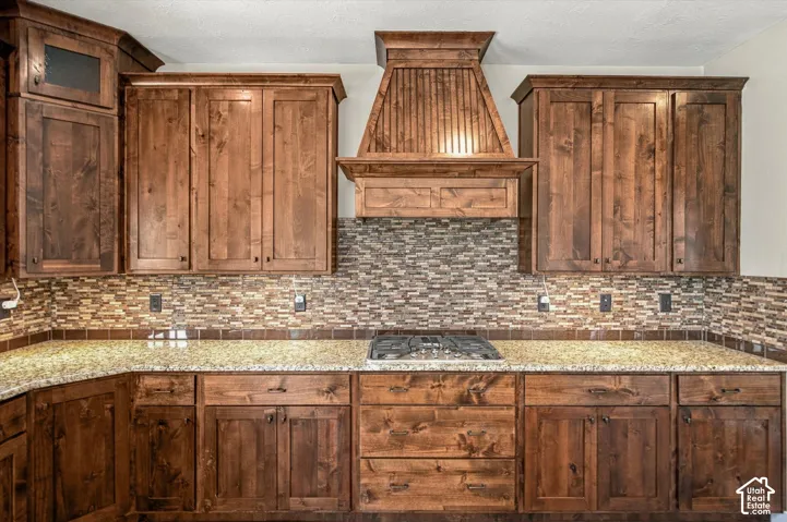 Custom cabinetry with warm wood finishes, integrated range hood, stone tile backsplash, stone-surface countertops, and built-in cooktop