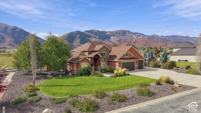 Multi-level exterior featuring a stone and siding facade, a three-car garage, expansive manicured lawn, tiered landscaping, and mountain backdrop