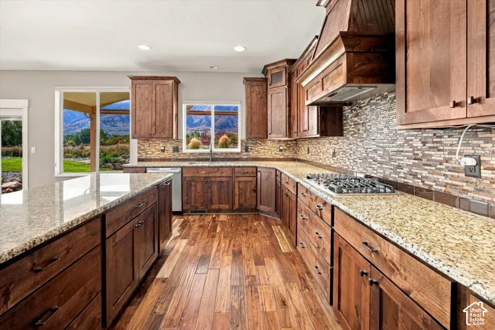 Kitchen featuring stained wood cabinetry, stone-tiled backsplash, expansive stone countertops, wood-finish flooring, and a built-in custom range hood