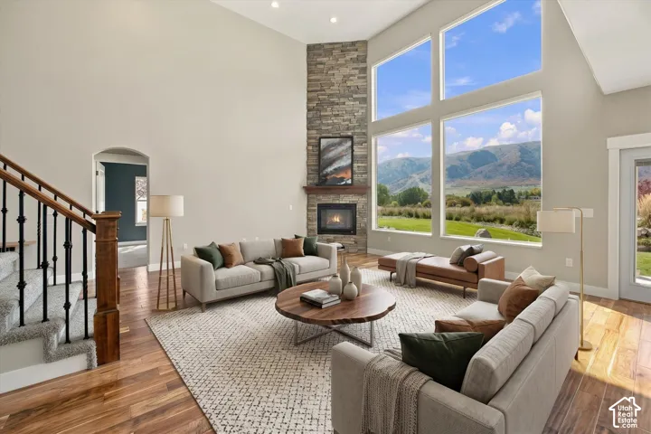High-ceiling living area featuring floor-to-ceiling windows with mountain scenery, stacked stone fireplace surround, wood-finish flooring, and staircase with metal balusters