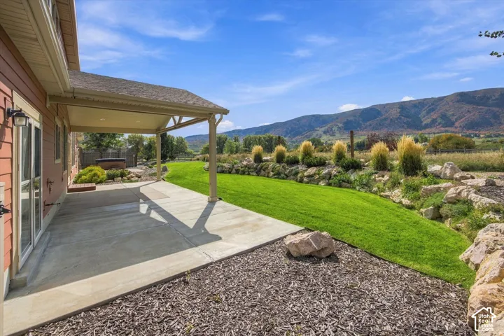 Covered concrete patio with recessed lighting, manicured lawn, tiered natural stone retaining wall, and mountain backdrop