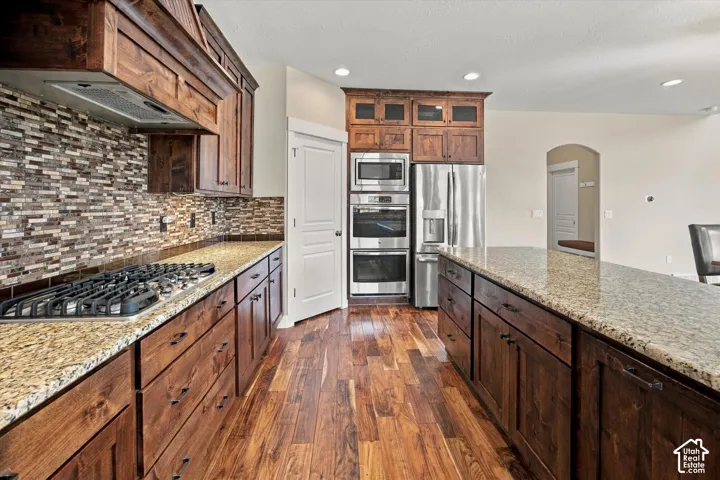 Kitchen featuring custom stained-wood cabinetry, stone countertops, a mosaic tile backsplash, stainless steel double wall ovens, and wood-finish flooring