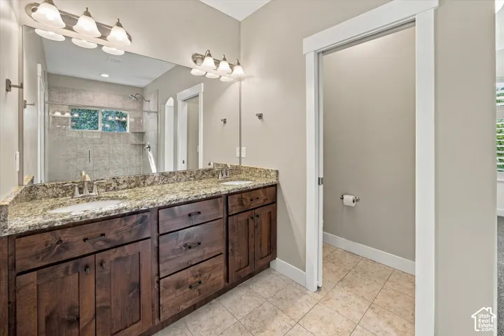 Dual-sink vanity featuring stone-finish countertops, dark wood-finish cabinetry, tile flooring, and wall-mounted multi-bulb lighting