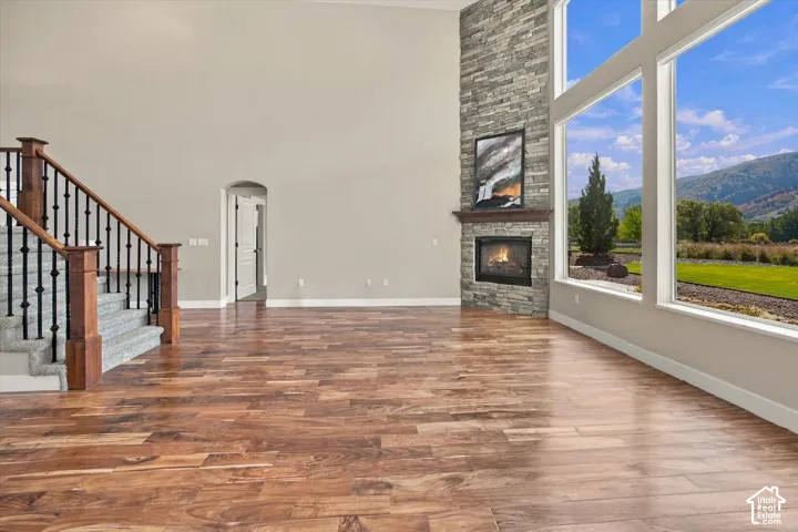 Expansive room with high ceilings, wood-finish flooring, floor-to-ceiling windows, stone-surround fireplace with a timber mantel, and an open staircase featuring wood newel posts and iron balusters